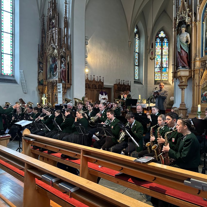 Ein Blechbläserensemble spielt in einer Kirche, ein Mann liest von einem Podium vor bunten Glasfenstern.