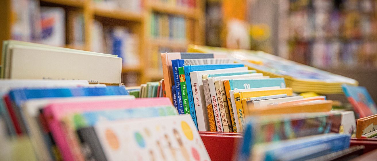 Ein Buchladen mit bunten Bücherreihen vorn, beleuchtet von Deckenlampen, und Holzregalen im Hintergrund.