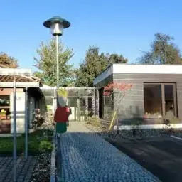 Eine Straßenlampe steht auf einem Gehweg, der zu einem modernen Gebäude führt. Das Gebäude hat Glasfenster und ein braunes Dach. Bäume und ein weiteres Gebäude sind im Hintergrund zu sehen.