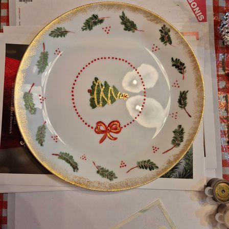 Ein weißer Keramikteller mit goldenem Rand, der einen Weihnachtsbaum, rote Punkte und grüne Blätter zeigt. Der Baum steht in der Mitte mit einem roten Band darum. Der Teller liegt auf einer weißen Oberfläche.