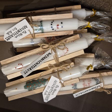 Eine Sammlung weißer Kerzen mit Weihnachtsdesigns ist in Plastik verpackt und auf Holzständern platziert. Jede Kerze hat einen dekorativen Anhänger mit festlichen Designs.