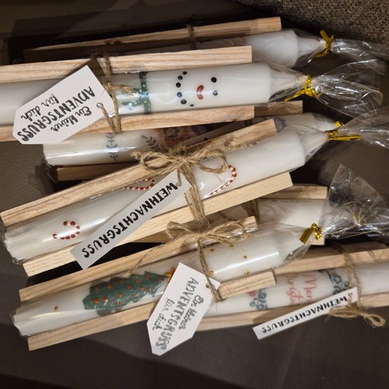 Eine Sammlung weißer Kerzen mit Weihnachtsdesigns ist in Plastik verpackt und auf Holzständern platziert. Jede Kerze hat einen dekorativen Anhänger mit festlichen Designs.