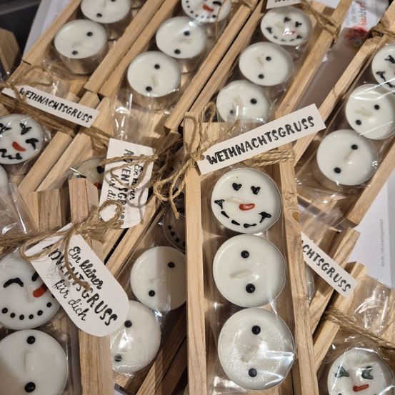 Mehrere Holzboxen mit Kerzen in Form von Schneemännern. Jede Kerze ist einzeln in Plastik verpackt und mit einer braunen Schnur gebunden. Auf den Schachteln steht 'Weihnachtsgruss'.
