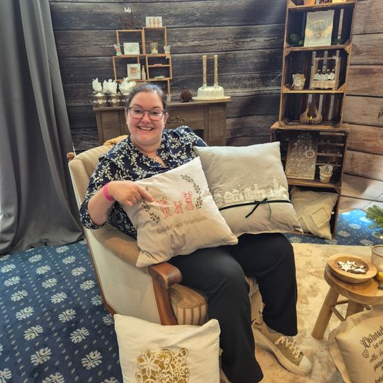 Eine Frau sitzt in einem Stuhl und hält ein Kissen mit Text und einem lächelnden Gesicht. Sie trägt eine Brille und ein Armband. Hinter ihr steht ein Holzregal mit Gegenständen. Der Boden ist mit einem Teppich mit Schneeflockenmuster bedeckt.