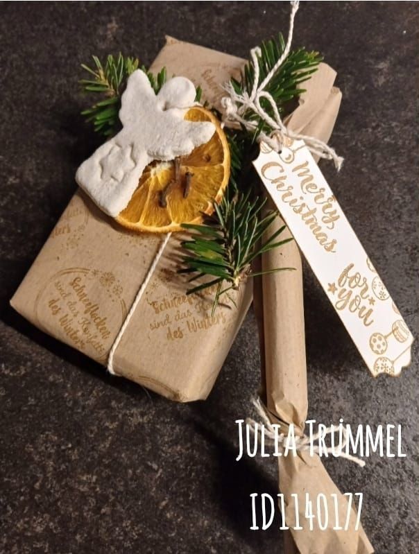 Ein verpacktes Geschenk mit einem Weihnachtsengel, Orangenscheibe und Tannenzapfen obenauf. Der Anhänger trägt 'Merry Christmas For You'. Geschaffen von Julia Trümmel. ID1140177.
