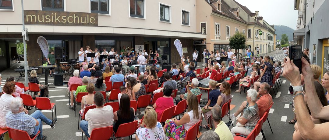 Ein Orchester spielt vor einem Gebäude mit dem Schild 'schule' vor einer Menschenmenge, die auf roten Stühlen sitzt.