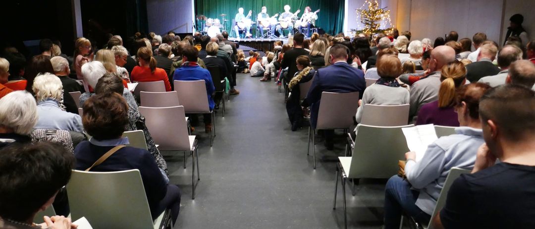 Eine Gruppe Musiker tritt auf der Bühne in einem Raum auf, in dem Menschen in Stühlen sitzen. Hinter den Musikern steht ein grüner Vorhang und rechts steht ein Weihnachtsbaum.