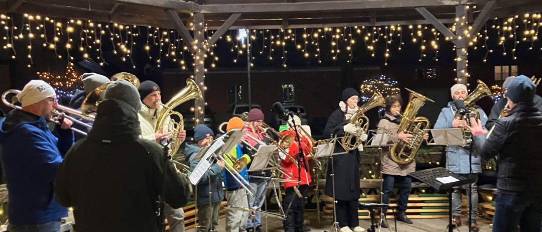 Ein Blechbläserensemble spielt unter einem mit Weihnachtslichtern geschmückten Pavillon, mit Musikern, die Tubas und Trompeten spielen, und Zuschauern, die die festliche Atmosphäre genießen.