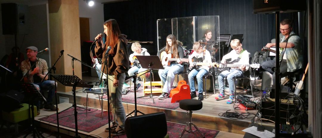 Eine Gruppe junger Menschen tritt auf der Bühne auf. Eine Frau singt in ein Mikrofon, während andere Gitarren spielen. Die Bühne ist mit Musikinstrumenten und bunten Stühlen geschmückt.