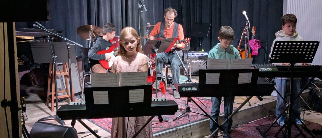 Ein junges Mädchen steht mit Notenblättern vor sich auf der Bühne, während zwei Jungen neben ihr stehen, einer mit einer Gitarre und der andere am Keyboard. Dahinter spielt ein Mann Gitarre und ein Schlagzeug ist aufgebaut.