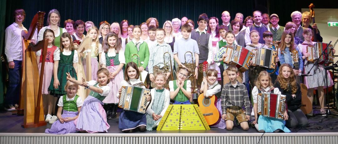 Eine Gruppe von Kindern und Erwachsenen auf einer Bühne mit Musikinstrumenten. Die Kinder halten Instrumente wie Akkordeon, Harfe und Trompete. Die Erwachsenen stehen hinter ihnen und schauen lächelnd auf die Kinder. Die Bühne hat einen schwarzen Vorhang und eine weiße Wand mit einem Lautsprecher.
