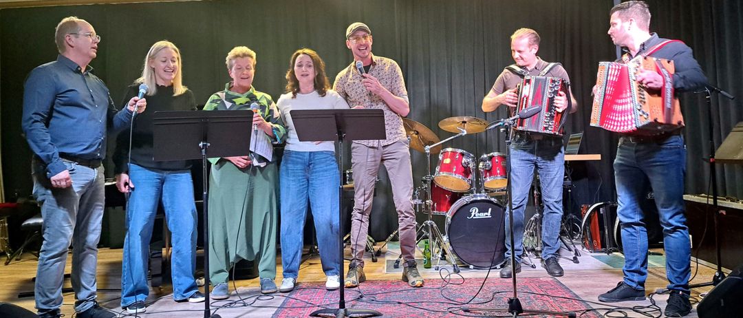 Eine Band tritt auf der Bühne auf, bestehend aus vier Mitgliedern. Zwei Frauen und ein Mann stehen vor einem Mikrofon, während der andere Mann ein Akkordeon spielt. Hinter ihnen steht ein Schlagzeug und eine Becken.
