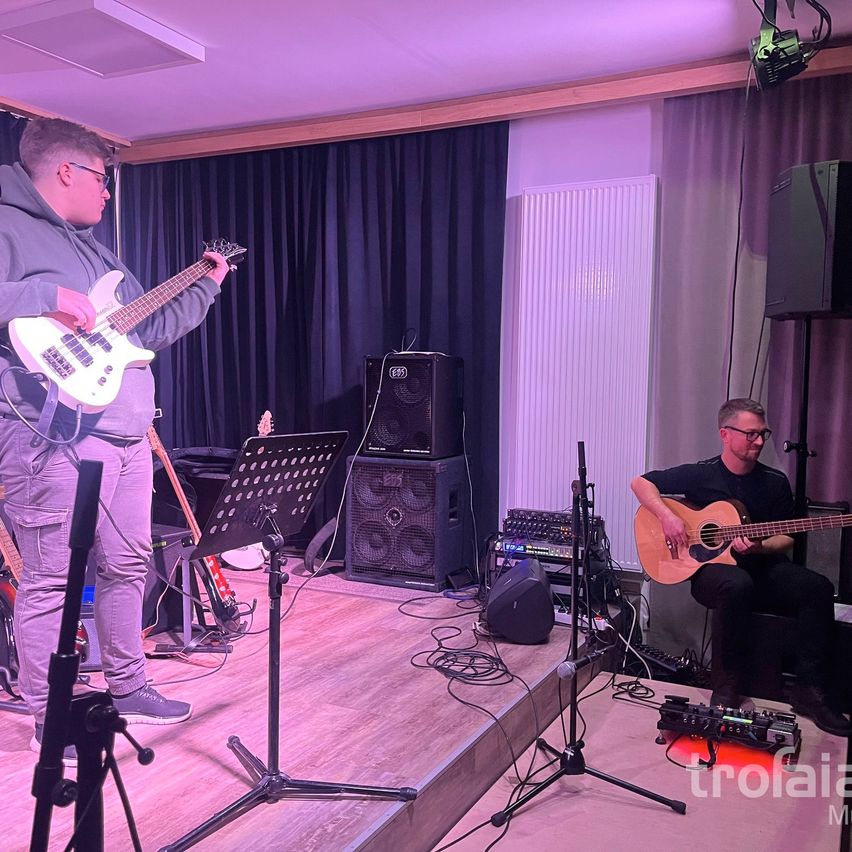 Zwei Männer spielen Gitarren auf einer Bühne. Einer steht, der andere sitzt. Sie sind von Ausrüstung umgeben. Ein Scheinwerfer und Vorhänge sind im Hintergrund.