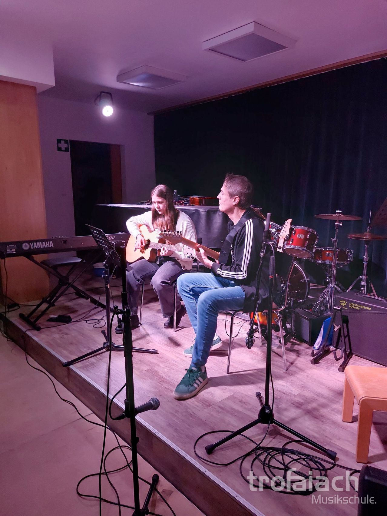 Zwei Personen sind auf einer Bühne, eine spielt Gitarre und die andere Schlagzeug, mit einem Yamaha-Piano und einem Yamaha-Keyboard im Hintergrund.