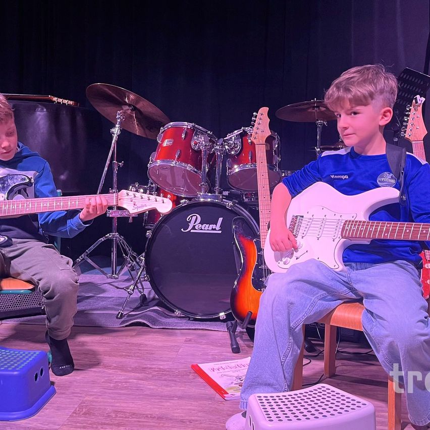 Zwei Jungen spielen Gitarren und Schlagzeug auf einer Bühne. Der Junge links spielt eine weiße Gitarre, während der Junge rechts ein schwarzes Schlagzeug spielt.