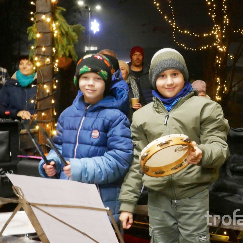 Zwei junge Jungen stehen bei Nacht draußen, einer hält einen Drumstick und der andere eine Trommel. Hinter ihnen steht eine Menschenmenge und Weihnachtslichter schmücken den Hintergrund.