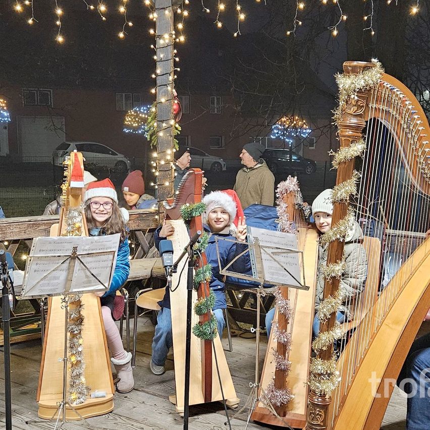 Eine Gruppe von Kindern mit Weihnachtsmützen spielt Harfen auf einer Bühne unter dekorativen Lichtern bei Nacht. Hinter ihnen beobachten zwei Erwachsene, und im Hintergrund sind Autos und Gebäude zu sehen.