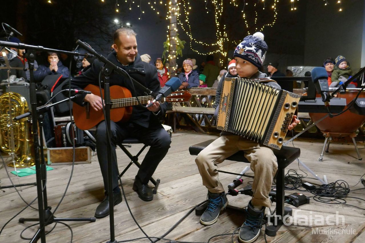 Ein Mann und ein Junge spielen nachts vor einem Publikum Musikinstrumente, mit Weihnachtslichtern im Hintergrund.