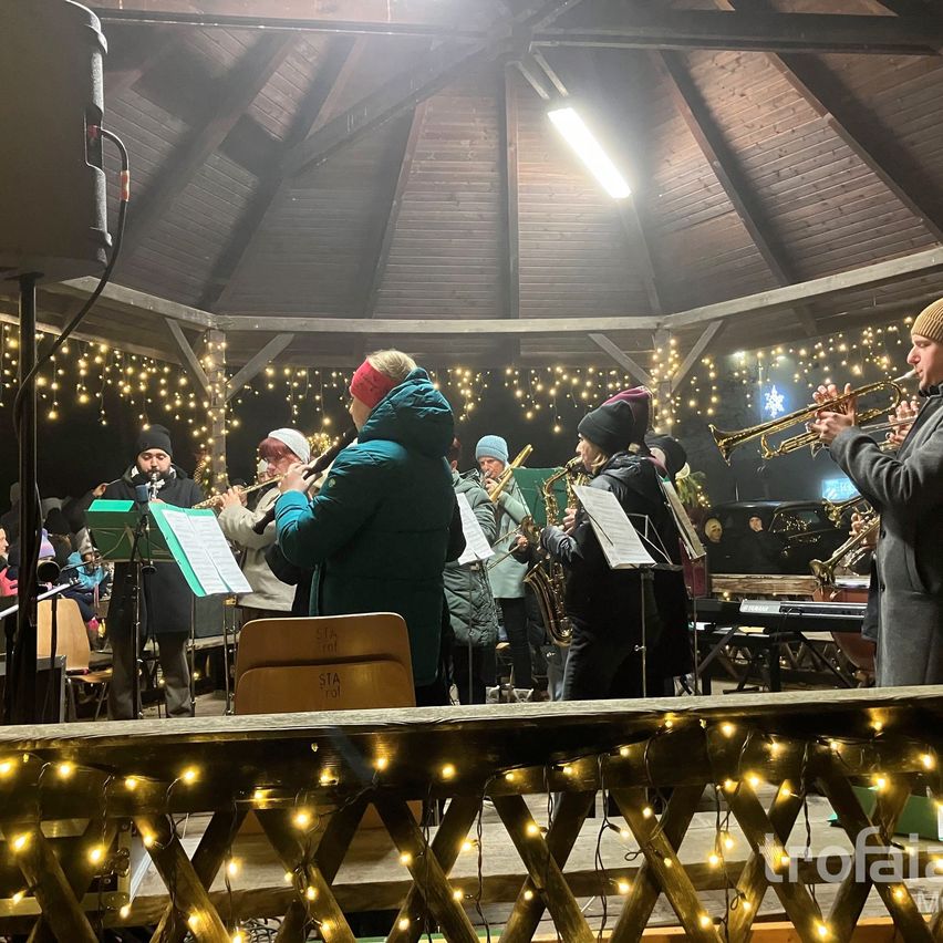 Eine Gruppe von Musikern spielt Trompeten und Flöten auf einer Bühne unter einem kuppelförmigen Dach mit Holzbalken und Lichtern.