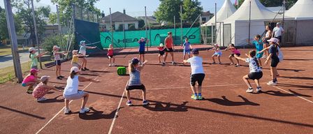 Eine Gruppe von Kindern und Erwachsenen spielt Tennis im Freien an einem sonnigen Tag, wobei eine Frau die Aktivität leitet. Ein grüner Banner mit dem Wort 'HEAD' steht hinter ihnen.
