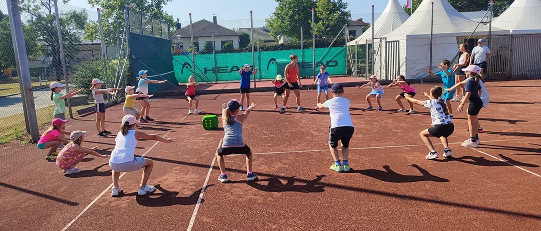 Eine Gruppe von Kindern und Erwachsenen spielt Tennis im Freien an einem sonnigen Tag, wobei eine Frau die Aktivität leitet. Ein grüner Banner mit dem Wort 'HEAD' steht hinter ihnen.