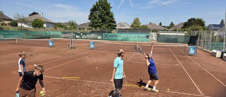Drei Personen sind auf einem Tennisplatz unter blauem Himmel. Einer bereitet sich auf das Aufschlag vor, ein anderer bereitet sich auf die Rückgabe vor, während der Dritte Bälle aus einem Korb holt.