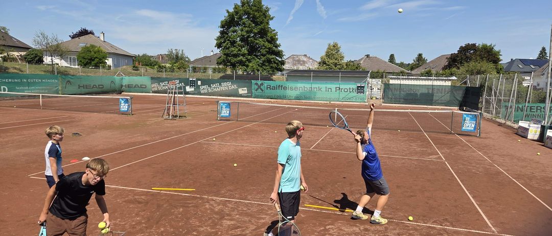 Drei Personen sind auf einem Tennisplatz unter blauem Himmel. Einer bereitet sich auf das Aufschlag vor, ein anderer bereitet sich auf die Rückgabe vor, während der Dritte Bälle aus einem Korb holt.