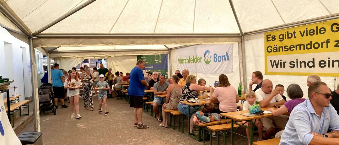 Eine Gruppe von Menschen speist in einem Zelt mit einem gelben Banner, das 'Es gibt Gänser' bewirbt. Einige sitzen, andere gehen. Ein Mann steht und spricht mit einem anderen. Tische sind mit Essen, Getränken und Hüten gedeckt.