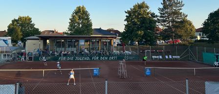 Ein Tennisspiel findet unter blauem Himmel statt, mit Spielern auf einem Sandplatz und Zuschauern im Hintergrund. Bäume und Gebäude sind in der Ferne sichtbar.