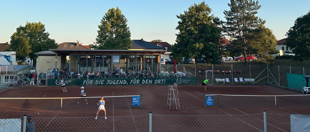 Ein Tennisspiel findet unter blauem Himmel statt, mit Spielern auf einem Sandplatz und Zuschauern im Hintergrund. Bäume und Gebäude sind in der Ferne sichtbar.