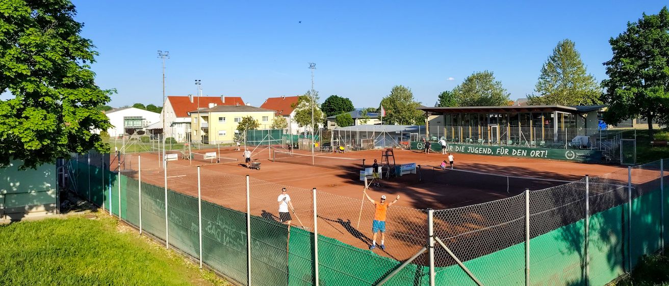 Ein Tennisplatz im Freien mit Spielern und einem Zaun, unter einem klaren Himmel. Ein Gebäude mit einem Schild 'Für die Jugend, für den Ort!' ist im Hintergrund.