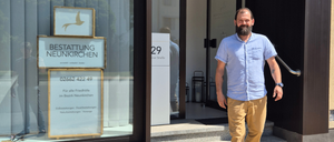 Ein Mann steht vor einem Geschäft in Glognitz, Österreich, mit einem Schild, das 'Betrieb Neunkirchen' und eine Telefonnummer anzeigt. Er lächelt und scheint Kunden willkommen zu heißen.