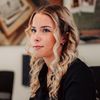 Eine Frau in einem schwarzen Oberteil arbeitet an einem Computer, mit einer Kollegin im Hintergrund. Ein Schreibtisch hält einen Computer, eine Tastatur und Papiere. Der Hintergrund umfasst ein gerahmtes Bild.