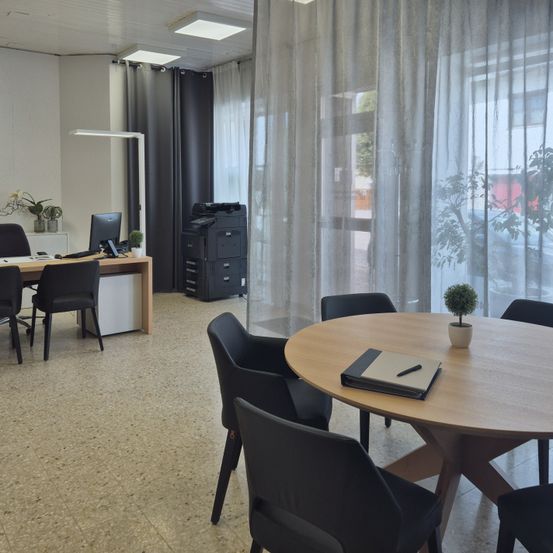 Ein Büro mit modernen Möbeln, einschließlich eines Schreibtischs mit Computer, Drucker und Topfpflanzen. Es gibt Stühle um einen runden Tisch mit einem Notizbuch und einem Stift. Der Raum hat weiße Wände, Deckenleuchten und ein Fenster mit weißen Vorhängen.