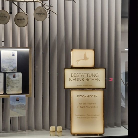 Ein Fensterdisplay mit Gedenktafeln, darunter eine mit einem Vogel und den Worten 'Bestattung Neunkirchen'. Es bietet Informationen zu Bestattungsdiensten für den Bereich Neunkirchen.