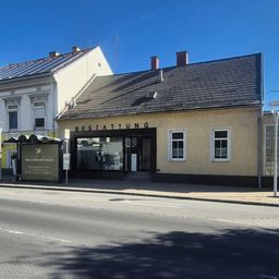 Ein Straßenblick auf ein Gebäude mit 'BESTATTUNG' in großen Buchstaben, begleitet von einem Bushaltestellenhäuschen mit einem Schild für 'Bestattungs Neunkirchen.' Das Gebäude hat ein graues Dach und gelbe Wände mit Fenstern.