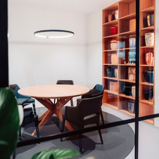 Ein runder Holztisch steht in der Mitte eines Raumes mit vier Stühlen. Ein Regal mit verschiedenen Vasen steht rechts. Eine kreisförmige Lampe hängt von der Decke.