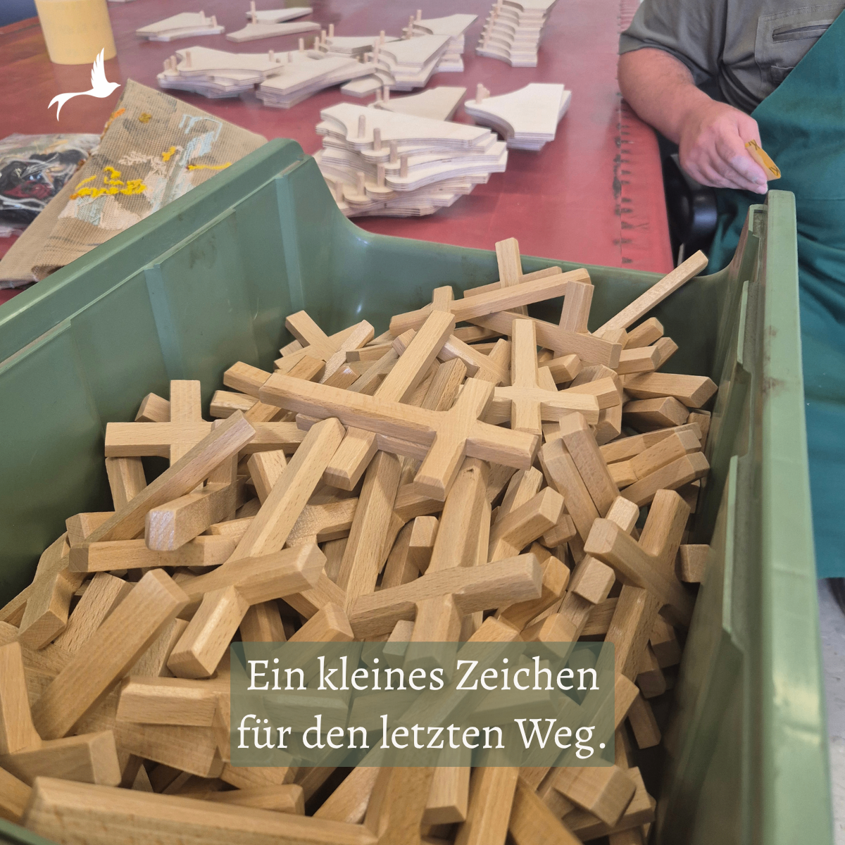 Ein grüner Aufbewahrungsbehälter gefüllt mit hölzernen Kreuzen, mit einer Person, die im Hintergrund an einem Holzstück arbeitet.