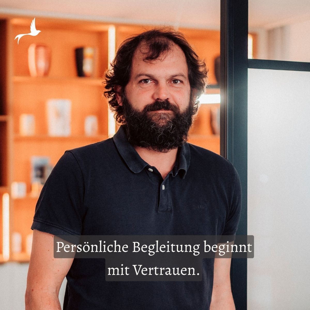A man with a beard stands in an office. Behind him is a wooden shelf with various items on it. He wears a dark blue polo shirt and looks ahead.