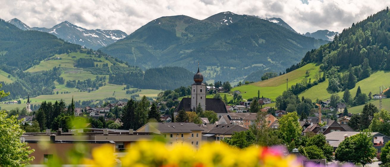 Eine Stadt in den Bergen mit einem Kirchturm, grünen Hügeln und einem bewölkten Himmel. Die Stadt hat viele Gebäude, Bäume und Blumen im Vordergrund.