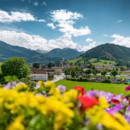 Ein malerisches Dorf in einem Tal, umgeben von üppigen grünen Hügeln und majestätischen Bergen unter einem blauen Himmel mit verstreuten Wolken.