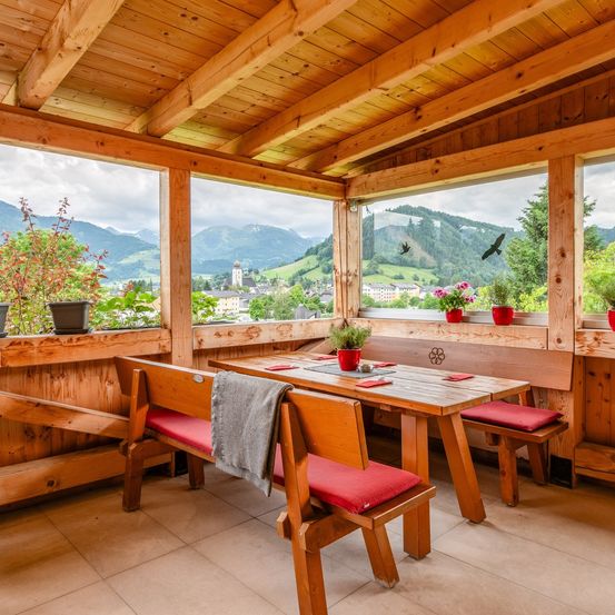 Eine Holzterrasse mit Tisch und Stühlen. Blumentöpfe und Blumen stehen auf der Fensterbank. Die Terrasse bietet Blick auf ein Dorf und Berge.