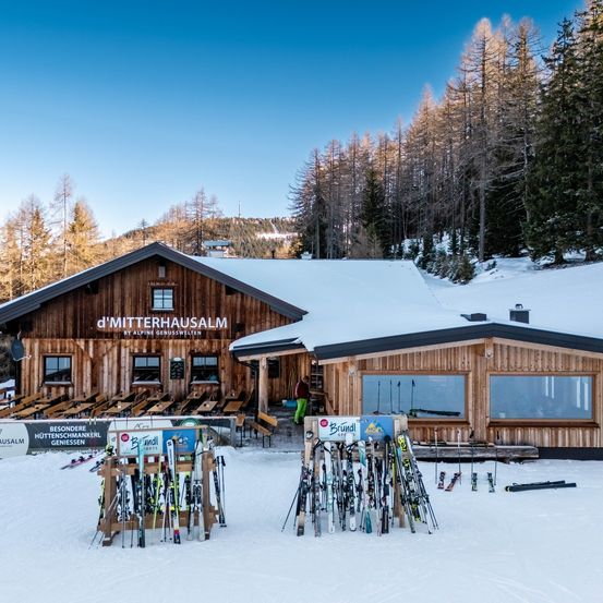 Eine Skihütte mit schneebedecktem Dach in einem Berggebiet. Vor der Hütte sind Skier und Stöcke ausgestellt. Eine Person in grüner Hose steht in der Nähe des Eingangs.