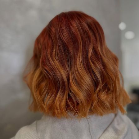 Ein Nahaufnahme des Rückens einer Person mit welligem, rötlich-braunem Haar. Das Haar ist in lockeren Wellen gestylt, und der Hintergrund ist unscharf.