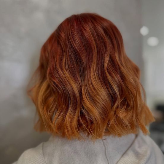 Ein Nahaufnahme des Rückens einer Person mit welligem, rötlich-braunem Haar. Das Haar ist in lockeren Wellen gestylt, und der Hintergrund ist unscharf.