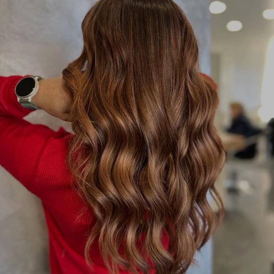Eine Frau mit langen, gewellten braunen Haaren in einem Salon gestyled, trägt einen roten Pullover und eine silberne Armbanduhr.