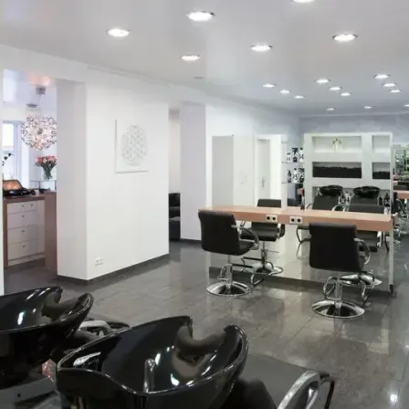 Ein leerer Salon mit weißen Wänden, schwarzen Stühlen und einem hölzernen Schreibtisch. Im Hintergrund sind eine Kronleuchter und eine Blumenvase zu sehen.