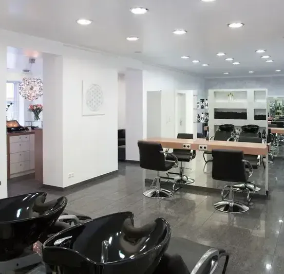 Ein leerer Salon mit weißen Wänden, schwarzen Stühlen und einem hölzernen Schreibtisch. Im Hintergrund sind eine Kronleuchter und eine Blumenvase zu sehen.
