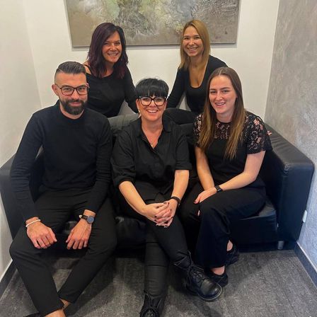 Fünf Menschen in schwarzer Kleidung sitzen auf einer Couch und lächeln für ein Foto. Der Mann links trägt eine Brille und eine Uhr.