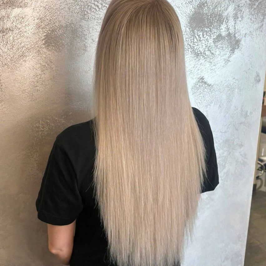 Eine Frau mit langen, glatten, hellblonden Haaren steht vor einer strukturierten Wand. Sie trägt ein schwarzes T-Shirt.